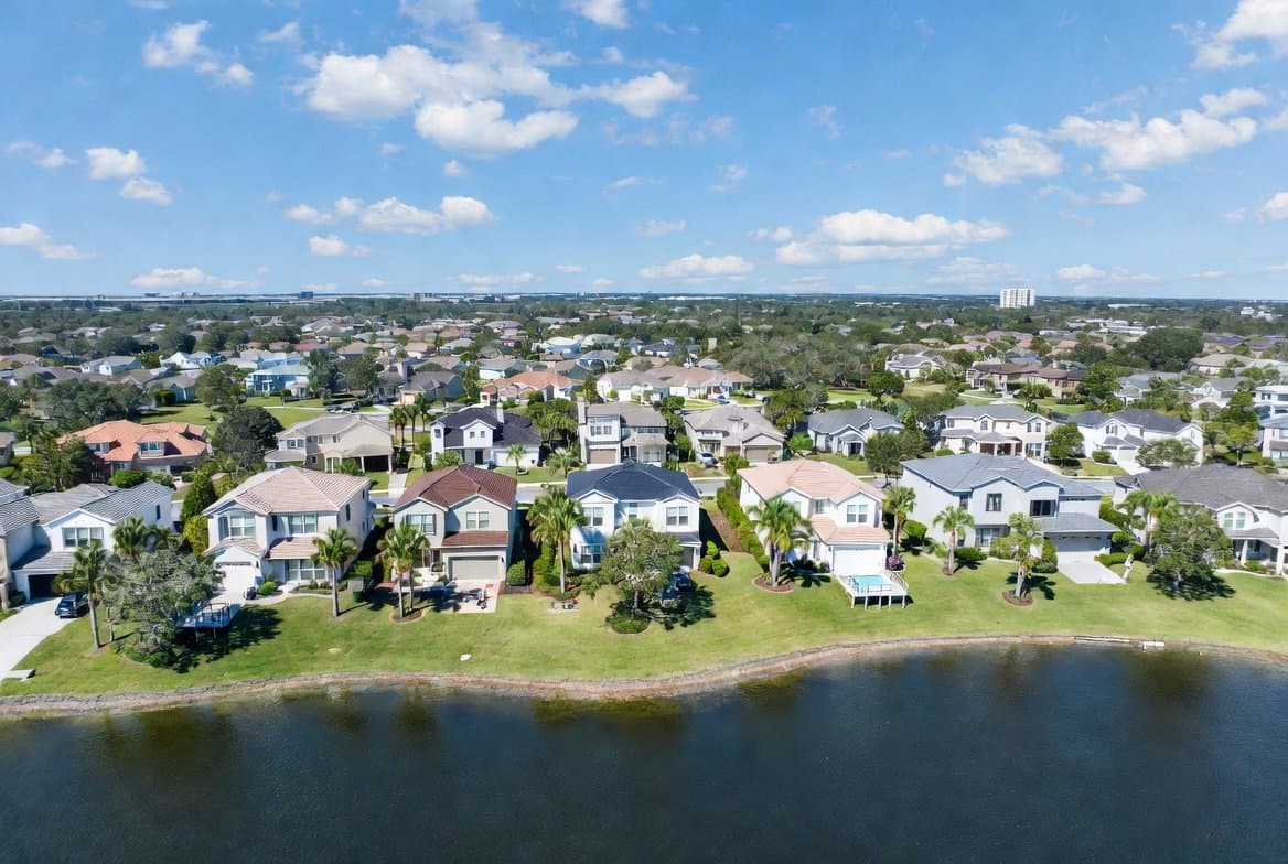 Aerial view of a premium Lutz, Florida neighborhood on a sunny day, showcasing modern single-family homes with lush lawns, palm trees, and a scenic lake in the foreground.