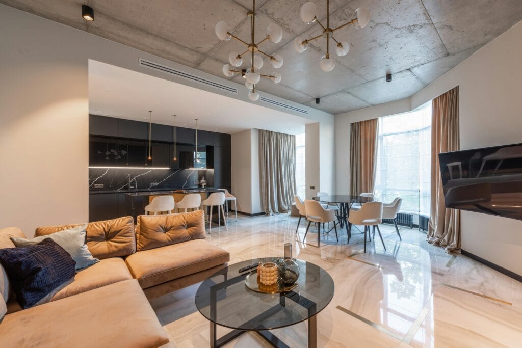 Interior of contemporary flat with sofa with pillows near kitchen with counter and round table with chairs near TV under chandelier near windows with curtains